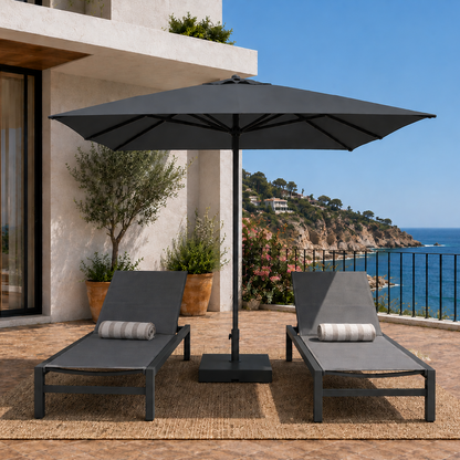 Two gray lounge chairs with a black umbrella on a patio overlooking the ocean.